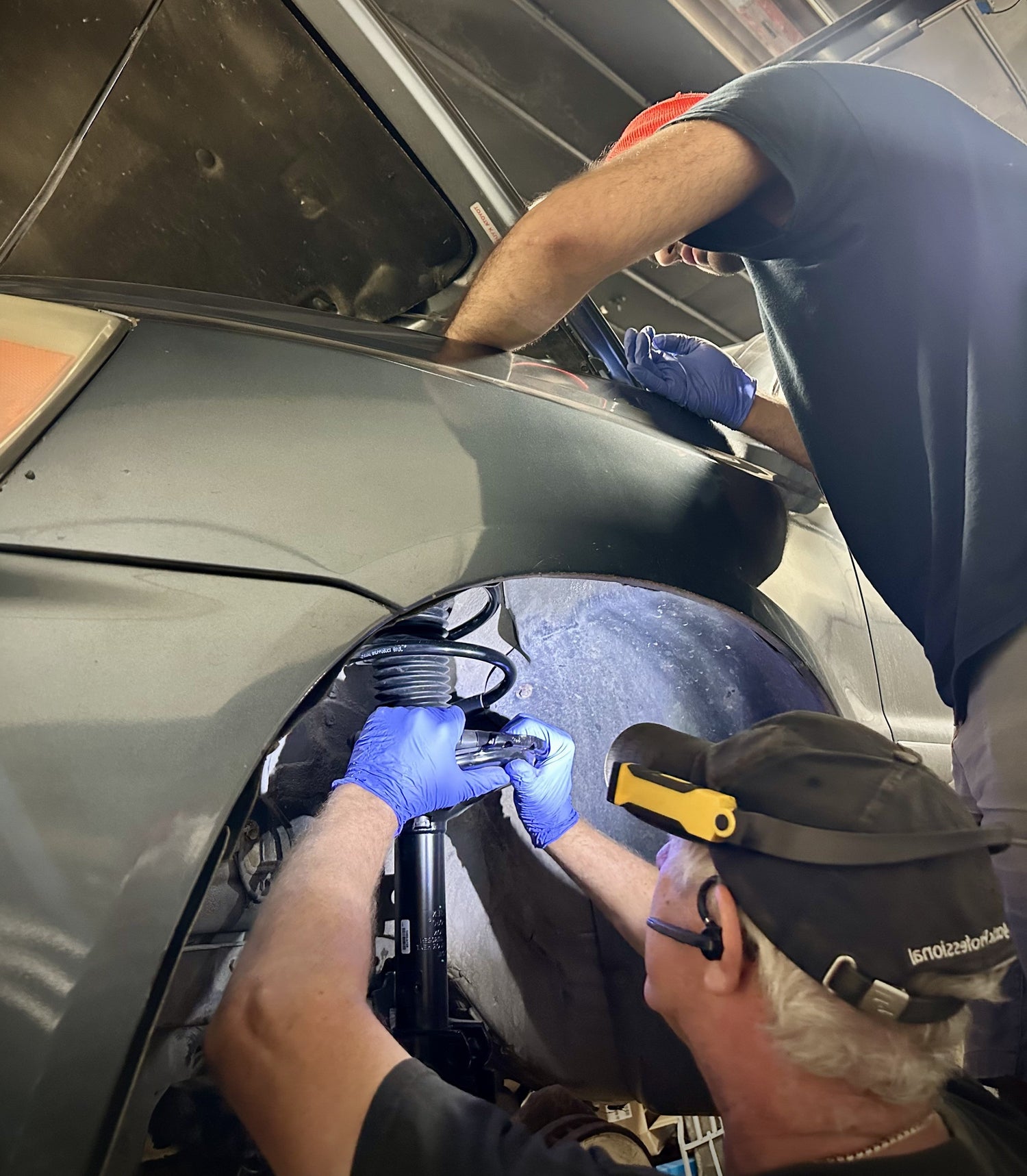 Two people working on a car with tools, one person wearing a black cap and gloves.